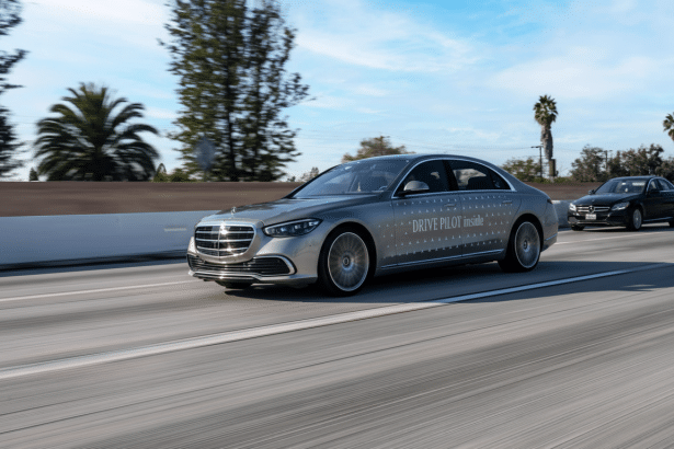 A silver Mercedes-Benz S-Class sedan with DRIVE PILOT inside text on its side , driving on a highway with palm trees in the background, resized to 16 :9 aspect ratio .
