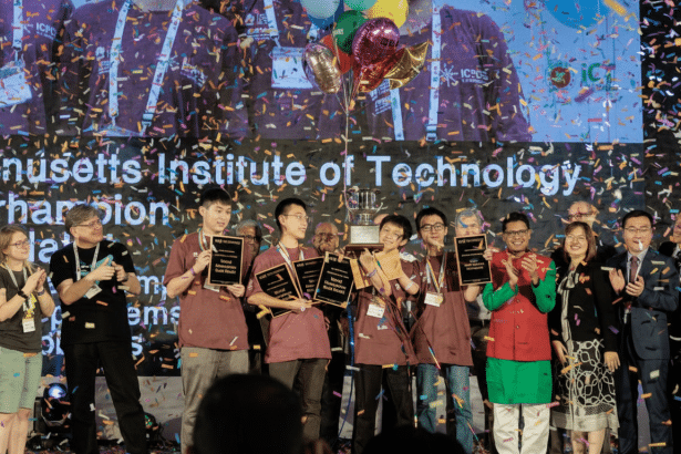 A group of students from MIT holding gold medals and a trophy on stage, surrounded by confetti, celebrating their championship win .