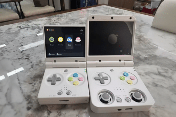 Two white handheld gaming devices , resembling a Nintendo DS , are placed side -by-side on a marbled table . The device on the left displays a user interface with app icons, while the device on the right has a black screen.