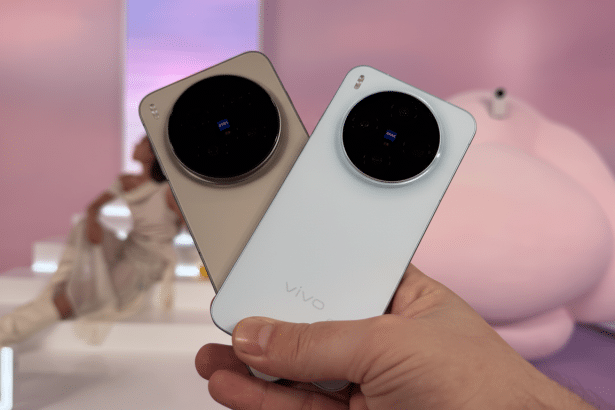 A hand holding two Vivo smartphones, one in a light beige color and the other in white, with large circular camera modules featuring Zeiss branding . A blurred background with pink and white elements and a person is visible. Filename : vivosmart phones16 9. png