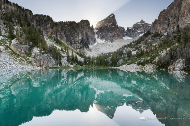 A stunning landscape image of a turquoise alpine lake perfectly reflecting the rugged mountain peaks and a bright sunbeam in the background.