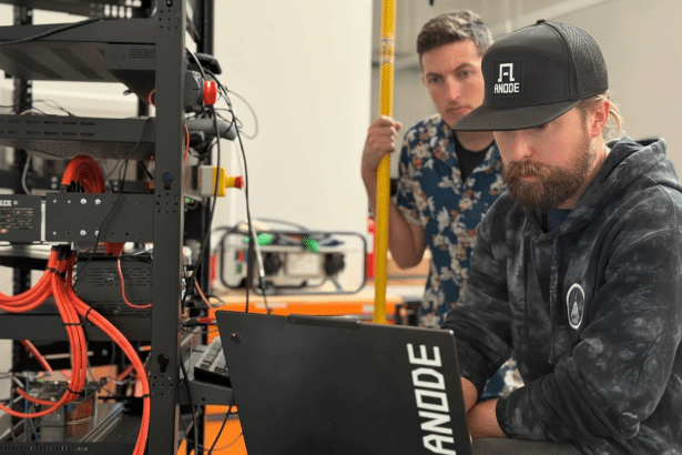 Two men working in a server room, with one focused on a laptop displaying the ANODE logo and the other observing.
