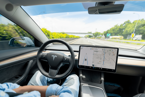 Interior view of a Tesla car with a driver's hands resting on their lap, showcasing the dashboard and a large central screen displaying navigation on a highway.