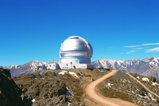 A large, silver -domed observatory sits atop a rocky hill, with a winding dirt road leading up to it. Snow -capped mountains are visible in the backgr