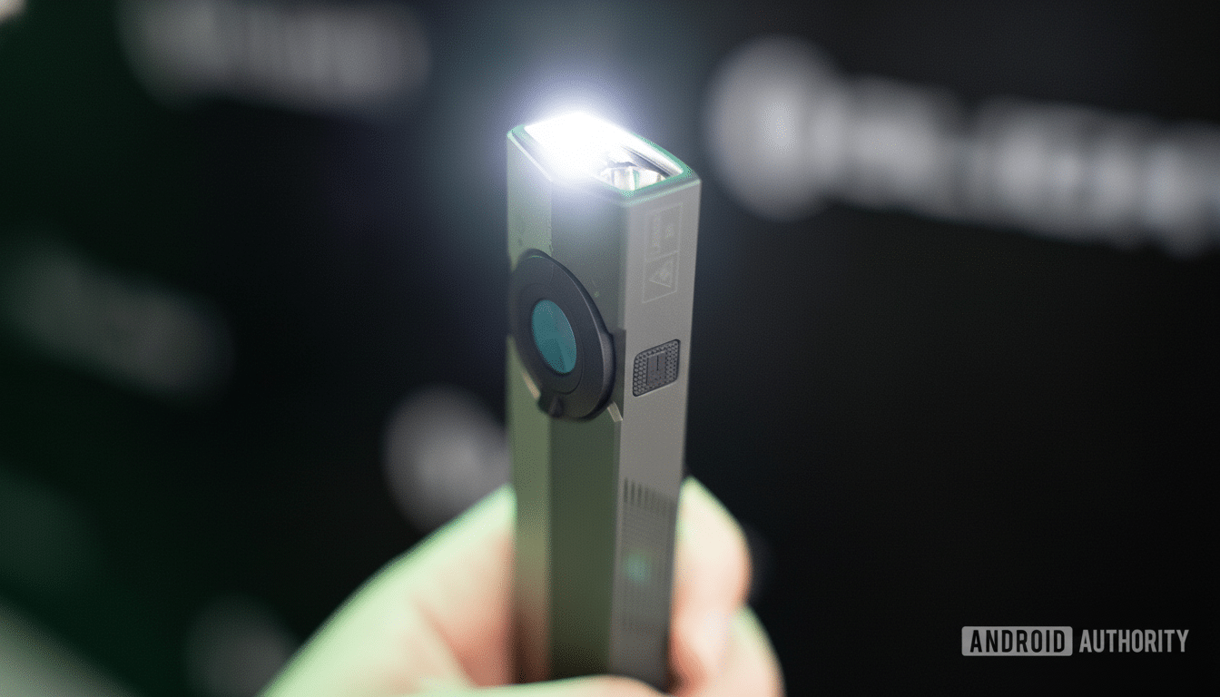 A hand holds a gray, rectangular device with a bright light illuminating from one end and a circular lens on its side.