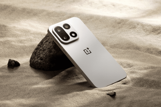 A professional shot of a silver OnePlus smartphone resting against a dark rock on sandy ground .