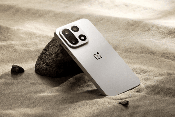A professional shot of a silver OnePlus smartphone resting against a dark rock on sandy ground, presented in a 1 6: 9 aspect ratio.