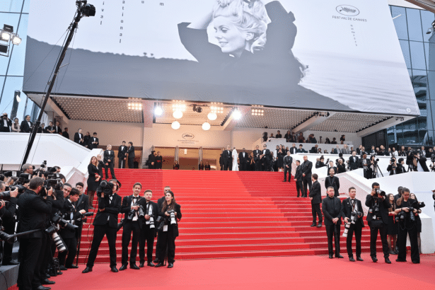 A wide shot of the red carpet event at the Cannes Film Festival, with a large black and white banner featuring an actress visible above the entrance.