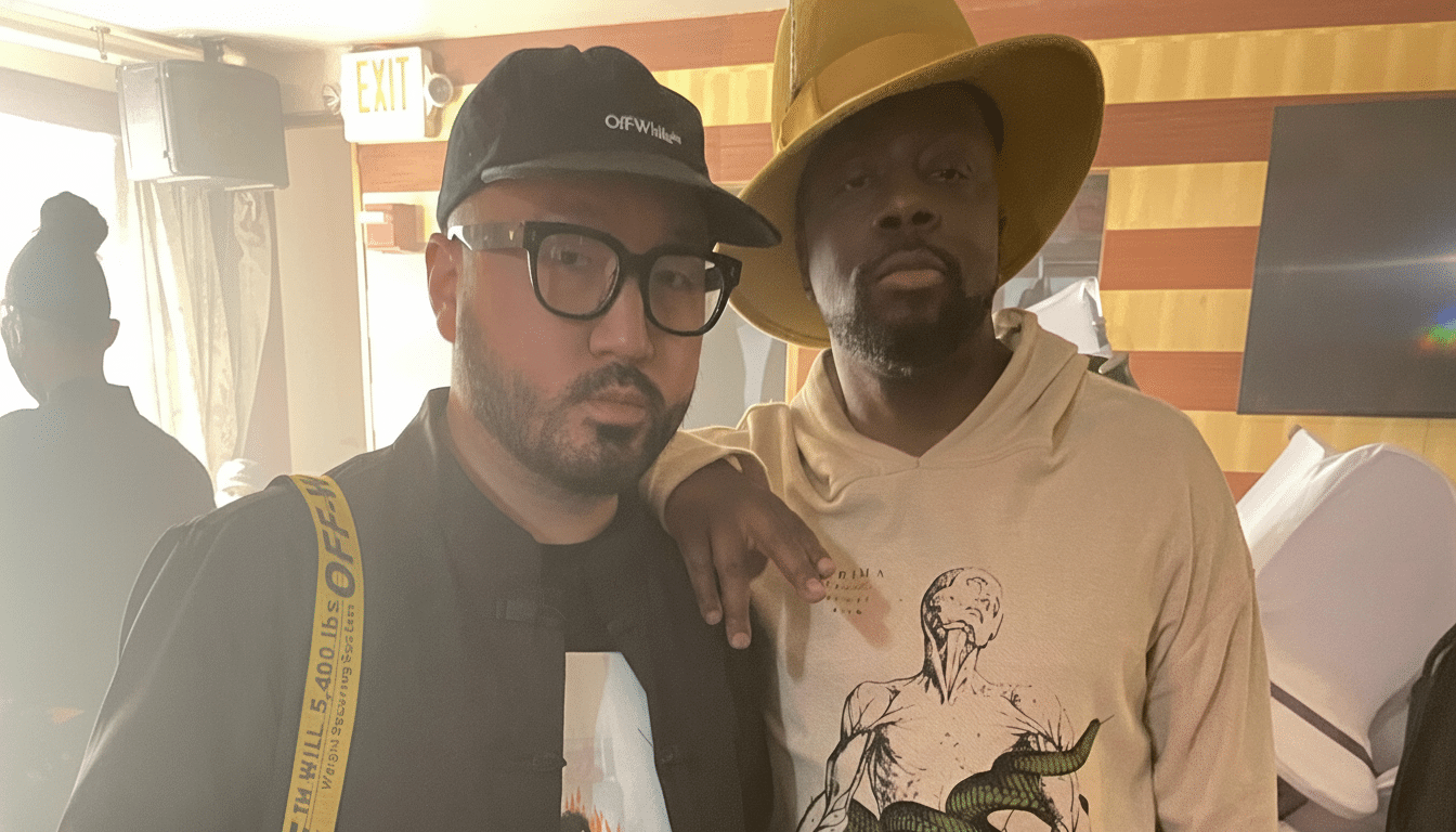 Two men, one wearing a black cap and glasses, the other in a wide -brimmed yellow hat and a graphic hoodie, stand together indoors.