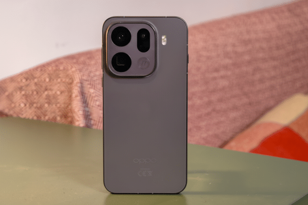 A grey smartphone with multiple cameras on its back, standing upright on a green surface with a blurry, patterned pink and red background.