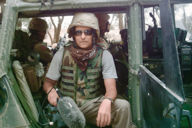 A person wearing a military helmet, sunglasses, a scarf , and a camouflage vest, holding a microphone, sitting in what appears to be a military vehicle . Another person in military gear is visible in the background.