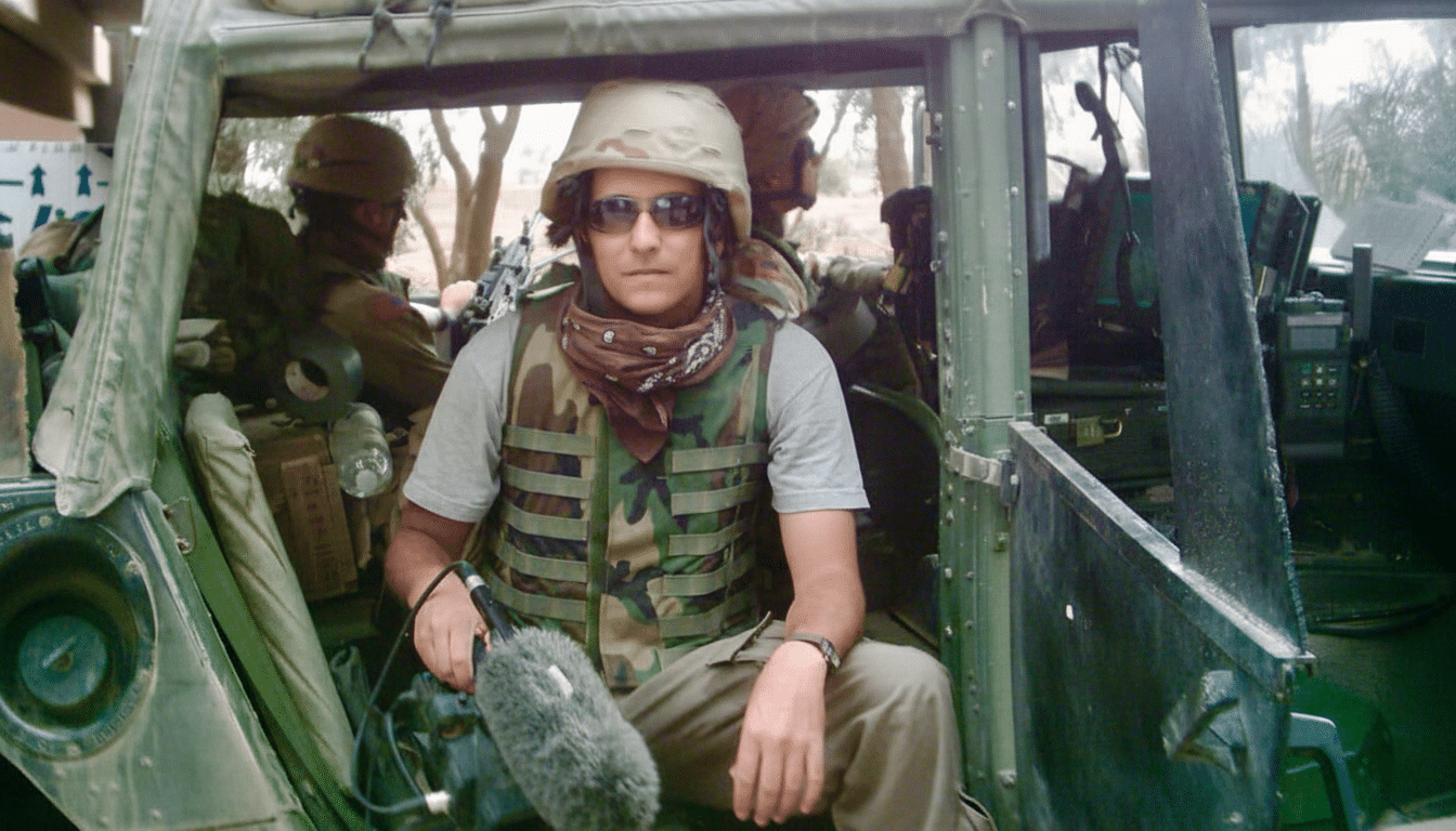 A person wearing a military helmet, sunglasses, a scarf , and a camouflage vest, holding a microphone, sitting in what appears to be a military vehicle . Another person in military gear is visible in the background.