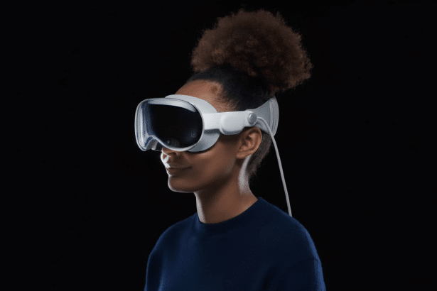 A person with dark, curly hair in a bun wearing white virtual reality goggles with a wire extending from the side , set against a dark background .