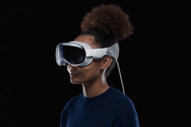 A person with dark, curly hair in a bun wearing white virtual reality goggles with a wire extending from the side, set against a dark background.