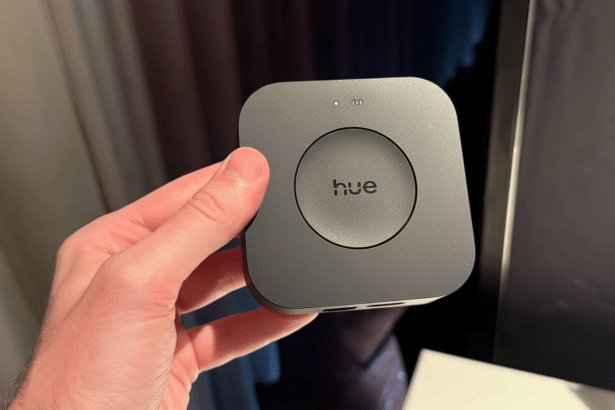A hand holding a dark gray Philips Hue Bridge device, featuring the hue logo in the center of a circular button. The background remains the original,