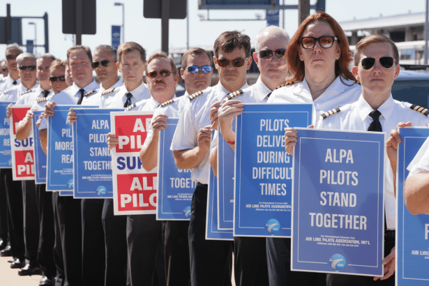 A line of airline pilots, some wearing sunglasses, holding blue and red signs with text supporting pilots rights and unity.