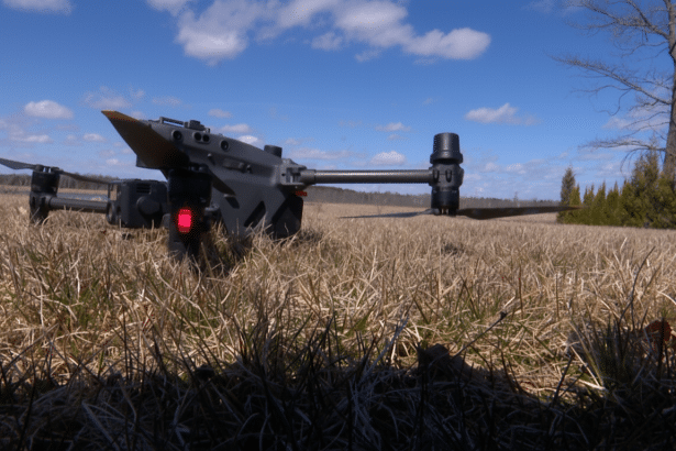 A professional drone with propellers resting in a field of dry grass under a blue sky with white clouds.
