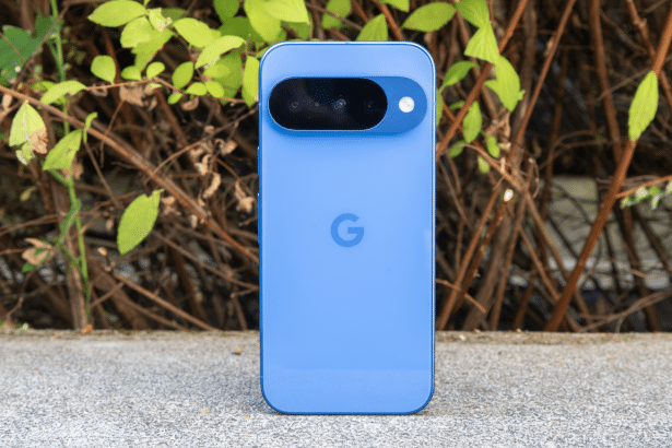 A blue Google Pixel 7 a smartphone is shown from the back, centered on a light gray concrete surface with a blurred background of green leaves and bro