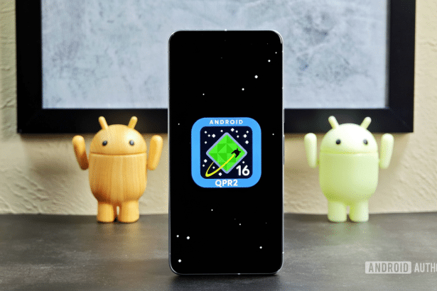 A smartphone displaying the Android 16 QPR 2 logo on a black screen, flanked by two Android figurines , set against a background with a framed picture. Filename : android1 6qpr 2smartphone . png