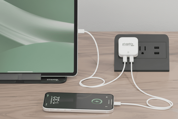 Plug able brand power adapter in a wall outlet, charging a tablet and a smartphone on a wooden surface.