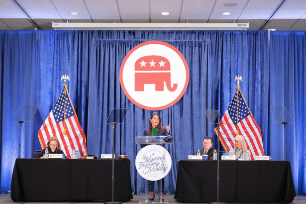 A woman speaks at a podium in front of a blue curtain with a Republican elephant logo, flanked by two American flags and seated panelists .