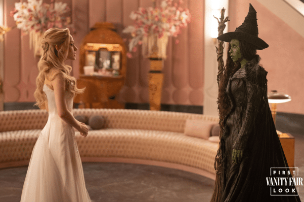 Two characters from Wicked, one in a white gown and the other as the green -skinned Wicked Witch , stand facing each other in a lavish room .