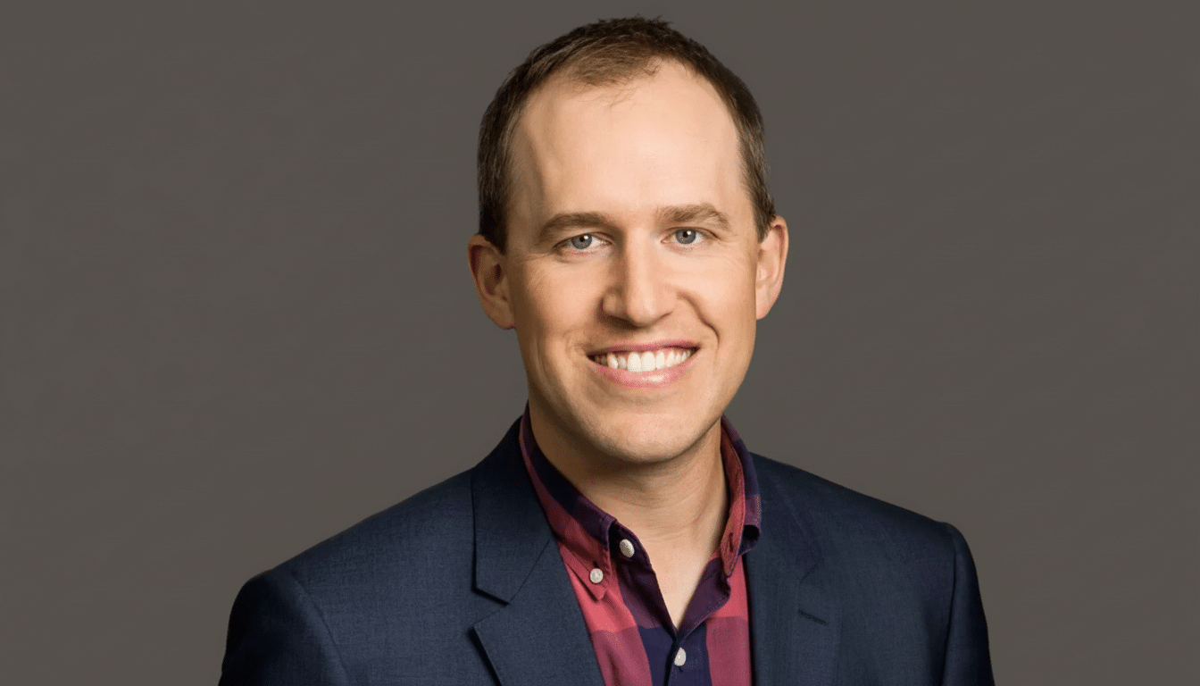 A professional head shot of a smiling man with short brown hair and blue eyes, wearing a blue suit jacket over a red and blue plaid shirt, set against