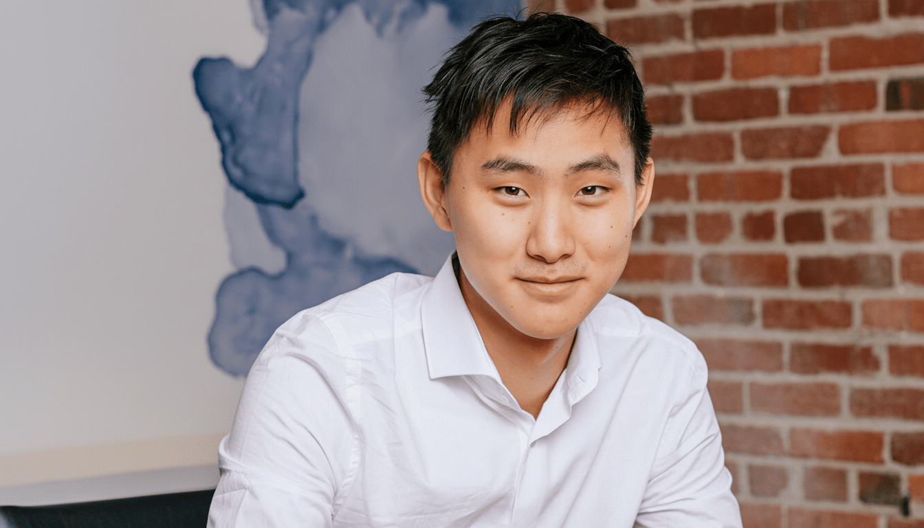 A professional head shot of a young East Asian man smiling, wearing a white button -up shirt, with a brick wall and a blue abstract painting in the ba