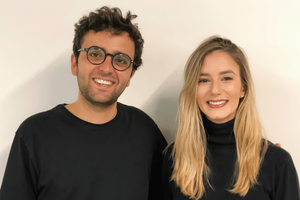 A professional head shot of a smiling man and woman , both dressed in black, standing against a plain white background. The man has dark curly hair and is wearing round glasses. The woman has long blonde hair.