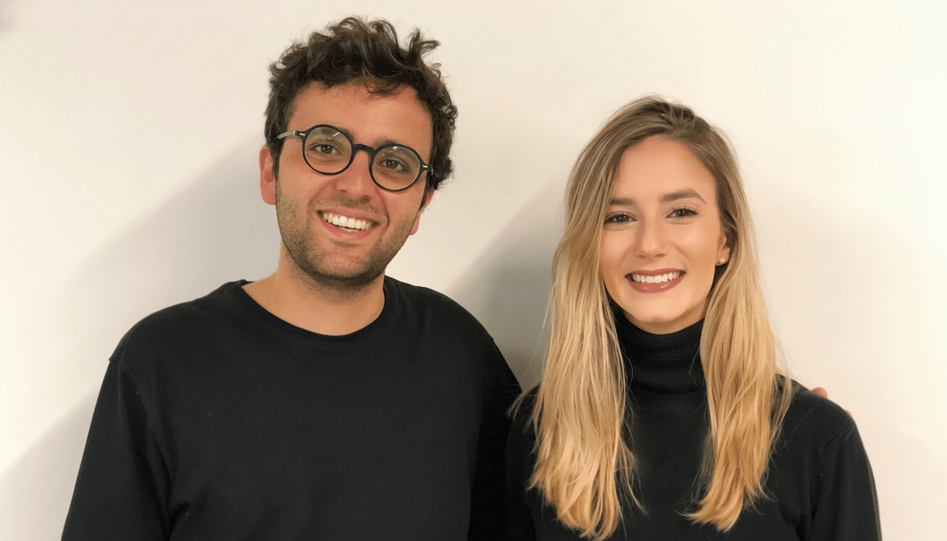 A professional head shot of a smiling man and woman , both dressed in black, standing against a plain white background. The man has dark curly hair and is wearing round glasses. The woman has long blonde hair.