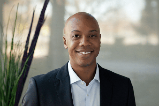 A professional portrait of a smiling bald man wearing a dark suit jacket and a light blue shirt , with a blurred office background featuring green and purple plant elements .