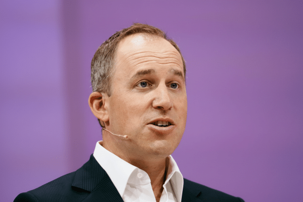 A man in a suit and white shirt with a lavalier microphone, speaking against a soft purple background.