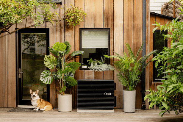 A cor gi sits next to a black Quilt home energy system, flanked by potted plants, against a modern wooden house.