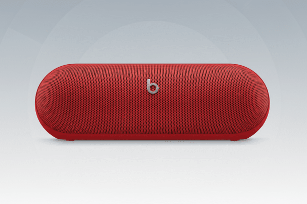 A red Beats Pill + portable Bluetooth speaker with the silver b logo centered on the front grille , set against a professional flat design background with a subtle gradient from light gray to white .