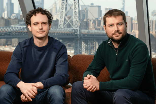 Two men sit on a brown sofa, looking at the camera, with a city skyline and bridge visible through a window behind them .