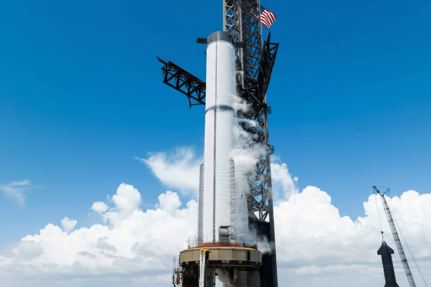 A large white rocket stands tall on a launchpad under a clear blue sky with white clouds. Smoke or vapor emanates from the rocket 's mid section, and an American flag is visible at the top of the launch tower structure. Another smaller rocket or structure is visible in the lower right.