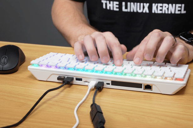 A person types on a white mechanical keyboard with RGB backlighting, connected by several cables, next to a black wireless mouse on a wooden desk.