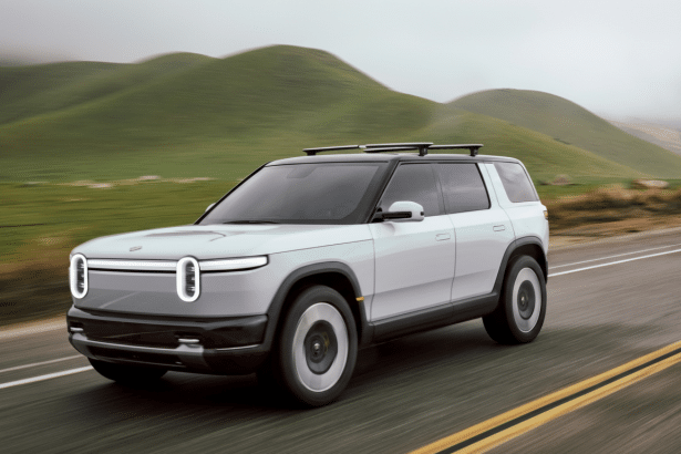 A professional shot of a light grey Rivian R 1S SUV driving on a road with green hills in the background, resized to a 16: 9 aspect ratio.