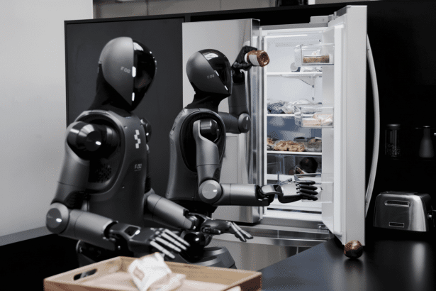 Two humanoid robots in a kitchen , one opening a refrigerator and holding a bottle, the other standing beside a counter.