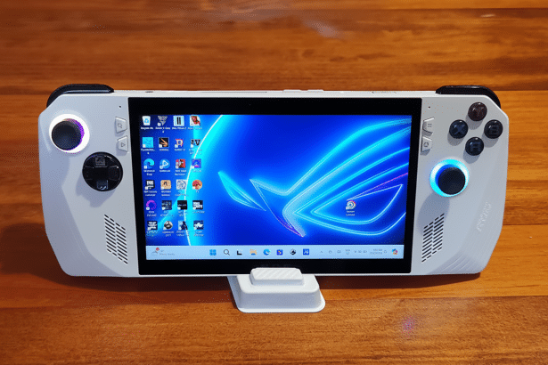 A white handheld gaming PC , the ROG Ally, displaying a blue desktop background with application icons, resting on a wooden surface.