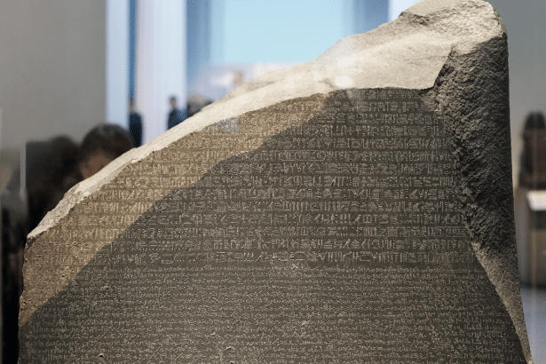 The Rosetta Stone, a dark gray gran odiorite ste le with ancient script , displayed in a museum setting with blurred human figures in the background. Filename : rosetta stonemuseum . png