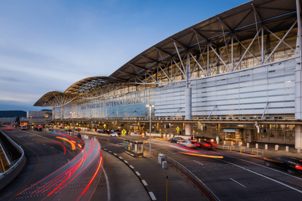 The exterior of San Francisco International Airport with light trails from cars at dusk, featuring modern architectural design and the airport ' s name visible on the terminal building. Filename : sanfranc iscointernational airportdusk .png