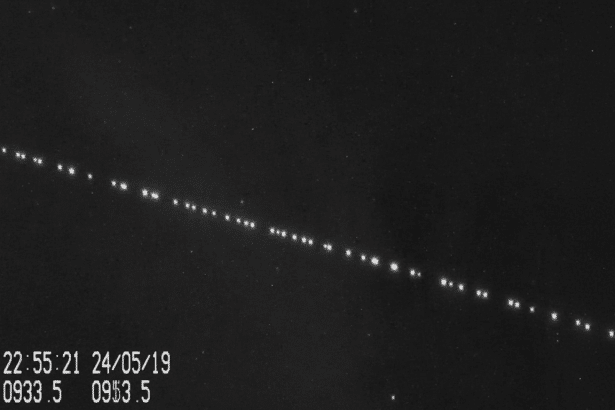 A professional , enhanced image showing a diagonal line of bright, evenly spaced lights against a dark background , likely indicating satellites in orbit. A timestamp and other numerical data are visible in the bottom left corner.