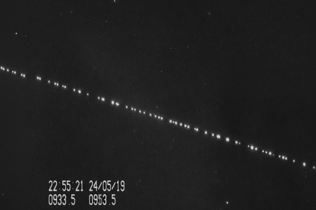 A black and white image showing a diagonal line of bright dots, likely satellites, against a dark starry sky, with a timestamp and coordinates in the