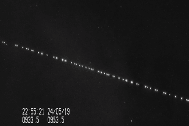 A black and white image showing a diagonal line of bright dots , likely satellites , against a dark starry sky. There is a timestamp and other data in the bottom left corner.