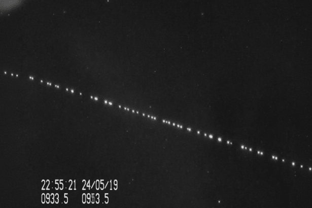 A black and white image showing a diagonal line of bright, evenly spaced dots against a dark background, likely indicating a satellite train or celestial observation . A timestamp and other numbers are visible in the bottom left corner .
