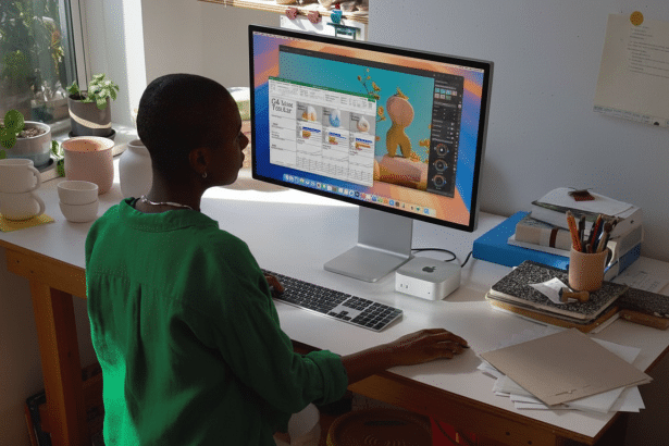 A person in a green shirt is seated at a desk, looking at a computer monitor displaying a spreadsheet application. The desk is cluttered with various office supplies , and natural light streams in from a window on the left.