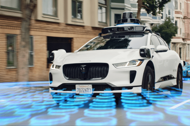 A white self -driving Jaguar I -Pace with a roof -mounted sensor array drives on a city street, with blue digital patterns emanating from beneath it .