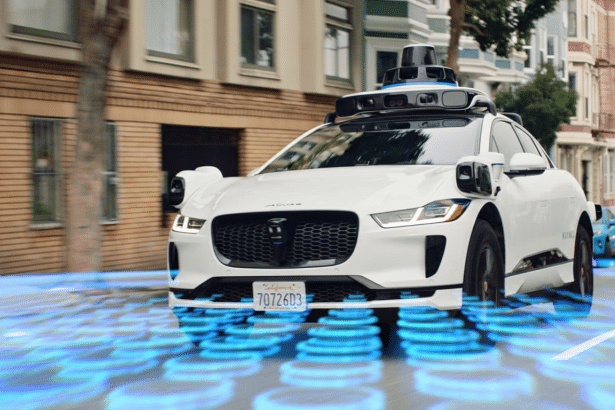 A white self -driving Jaguar I -Pace with advanced sensors on its roof and sides , driving on a city street with a blue digital overlay on the road.