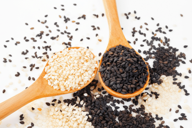 Two wooden spoons, one filled with white sesame seeds and the other with black sesame seeds, arranged on a white surface with scattered seeds. Filename : sesameseeds spoons. png
